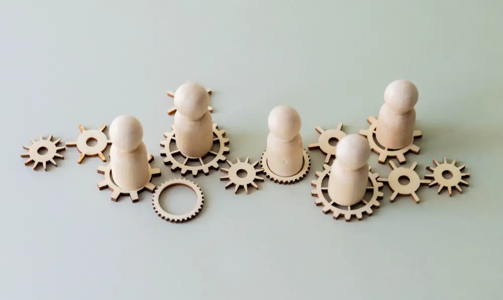 Bilden visar tr&auml;figurer som st&aring;r p&aring; kugghjul. Bilden har ett dekorativt syfte f&ouml;r kursen Utveckla processledning som h&aring;lls av Trivector LogiQ.